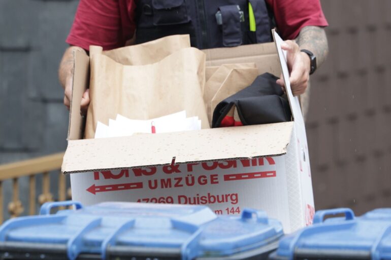 Ein Polizist trägt nach einer Wohnungsdurchsuchung einen Karton mit sichergestellten Gegenständen.