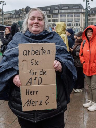Eine Demonstrantin in Hamburg stellt auf einem Pappschild eine Frage an Bundeskanzler Friedrich Merz (CDU).
