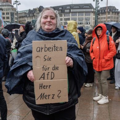 Eine Demonstrantin in Hamburg stellt auf einem Pappschild eine Frage an Bundeskanzler Friedrich Merz (CDU).