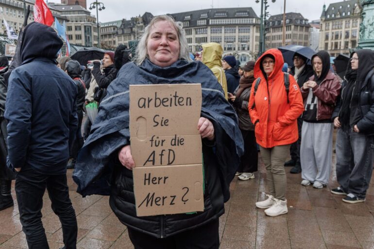 Eine Demonstrantin in Hamburg stellt auf einem Pappschild eine Frage an Bundeskanzler Friedrich Merz (CDU).