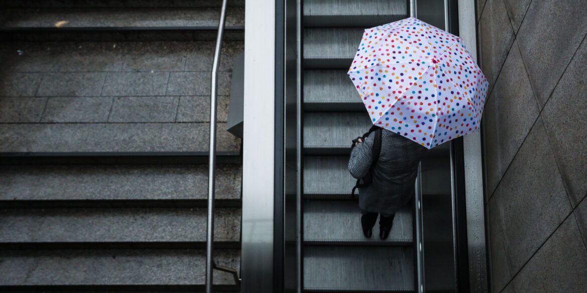 Eine Frau fährt mit ihrem Regenschirm eine Rolltreppe hinauf. Das Wetter soll in der Region Stuttgart in den nächsten Tagen wechselhaft bleiben.