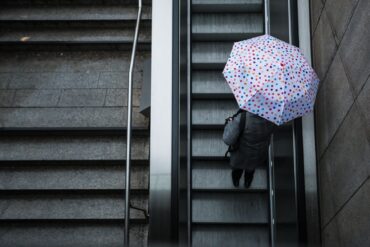 Eine Frau fährt mit ihrem Regenschirm eine Rolltreppe hinauf. Das Wetter soll in der Region Stuttgart in den nächsten Tagen wechselhaft bleiben.