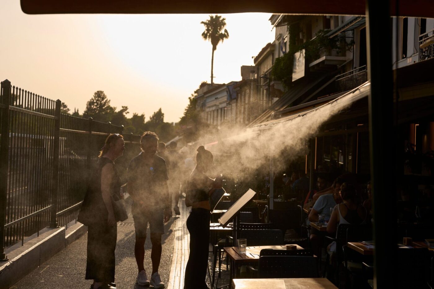 Ein Touristenpaar geht an einer Sprühanlage vorbei, während ein Restaurantmitarbeiter sie im Athener Stadtteil Monastiraki zu ihrem Tisch führt.