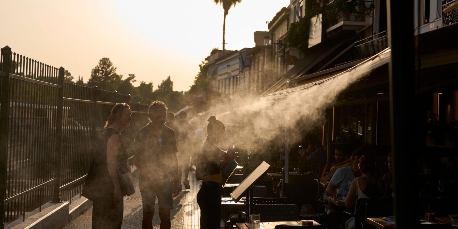 Ein Touristenpaar geht an einer Sprühanlage vorbei, während ein Restaurantmitarbeiter sie im Athener Stadtteil Monastiraki zu ihrem Tisch führt.