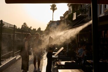 Ein Touristenpaar geht an einer Sprühanlage vorbei, während ein Restaurantmitarbeiter sie im Athener Stadtteil Monastiraki zu ihrem Tisch führt.