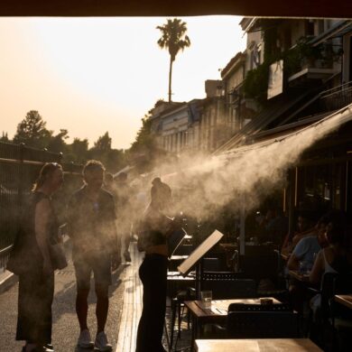 Ein Touristenpaar geht an einer Sprühanlage vorbei, während ein Restaurantmitarbeiter sie im Athener Stadtteil Monastiraki zu ihrem Tisch führt.