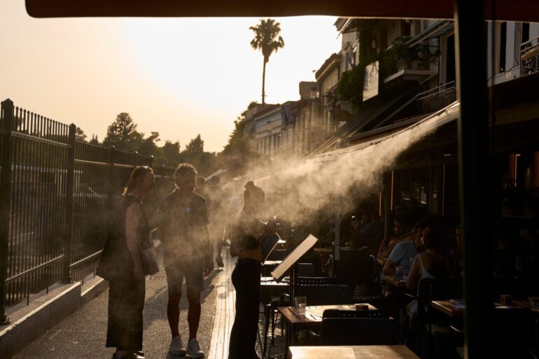 Ein Touristenpaar geht an einer Sprühanlage vorbei, während ein Restaurantmitarbeiter sie im Athener Stadtteil Monastiraki zu ihrem Tisch führt.