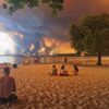 Strandbesucher sitzen am Strand von Moutchic in Lacanau im Südwesten Frankreichs, während Rauchwolken von einem Waldbrand in den Himmel aufsteigen.