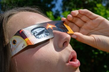 Eine junge Frau schaut durch eine Sonnenfinsternisbrille und betrachtet die Mittagssonne.