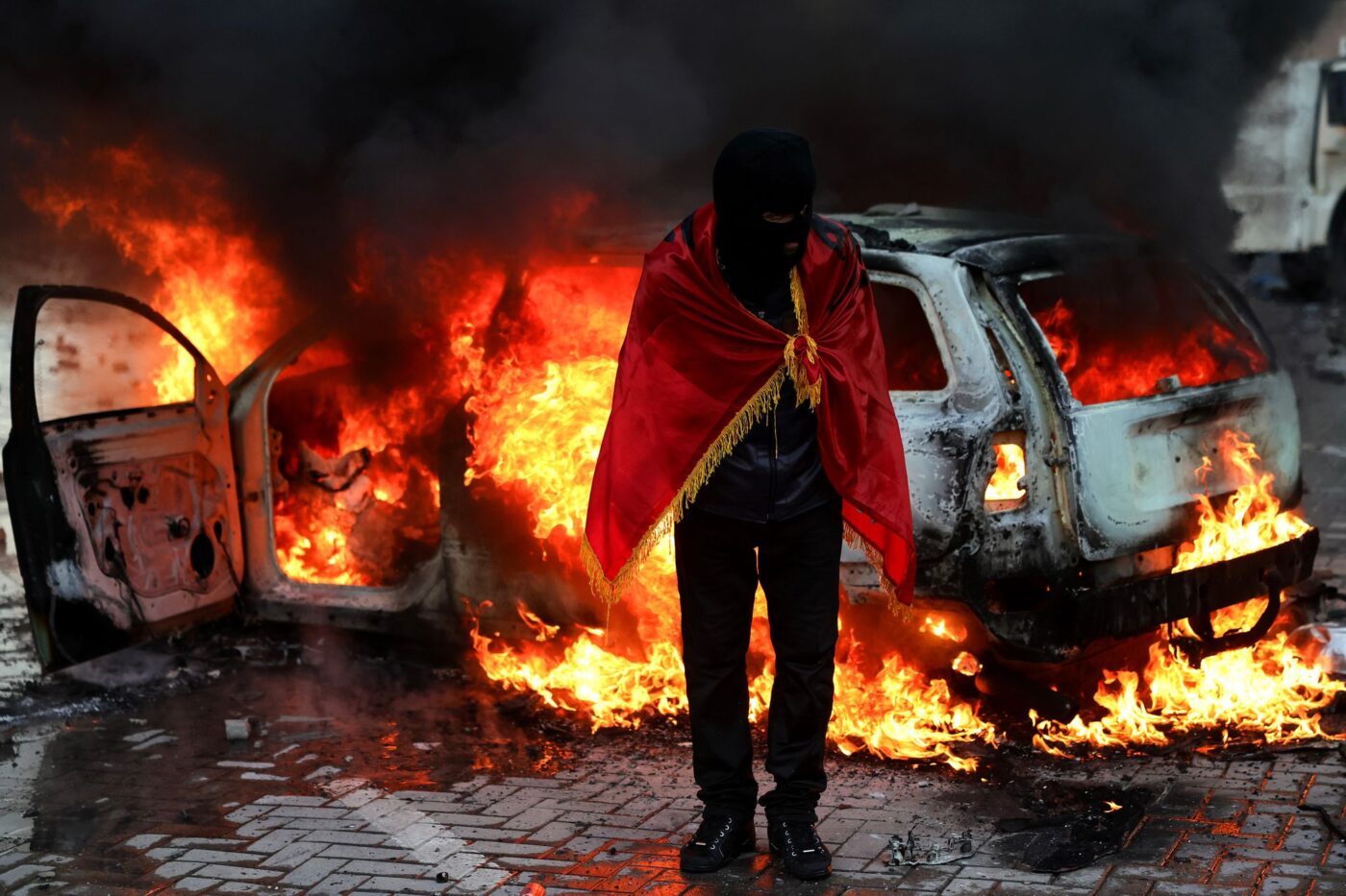 Ein Demonstrant, der sich in eine albanische Flagge gehüllt hat, steht während einer regierungskritischen Demonstration in Tirana vor dem Büro des Ministerpräsidenten vor einem brennenden Auto.